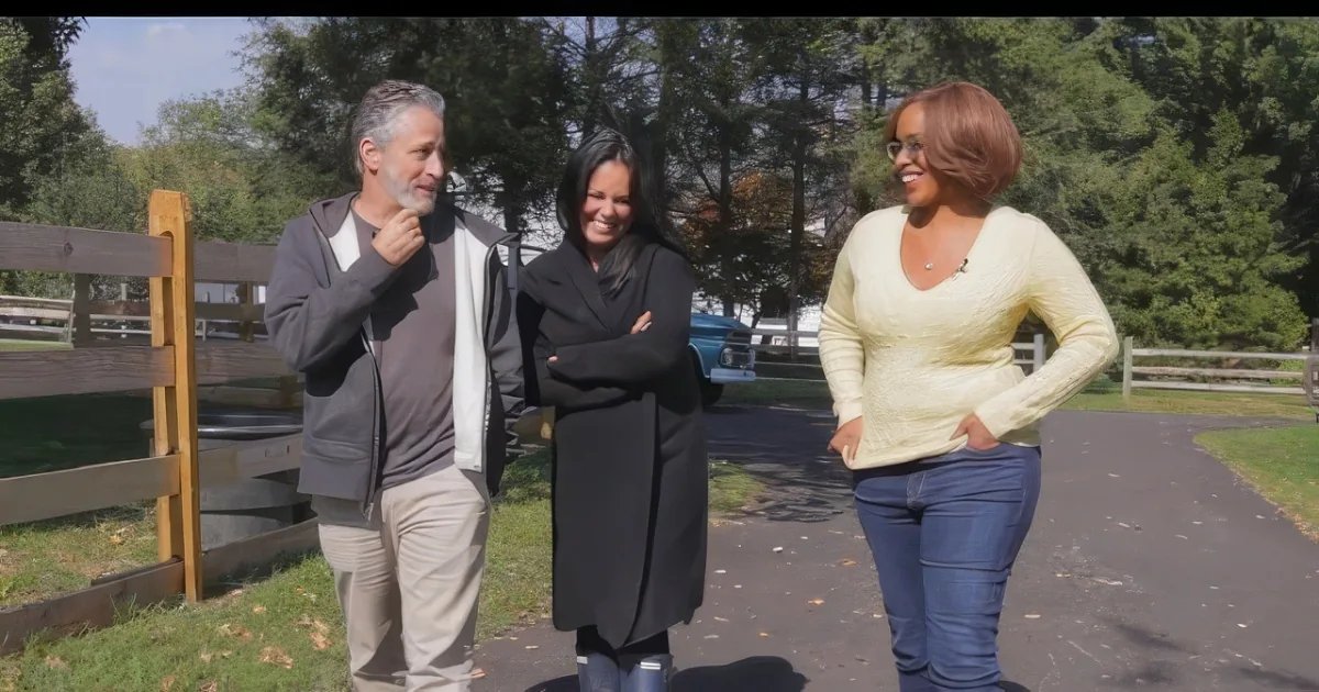 Three adults stand outdoors, surrounded by trees and grass; a paved path suggests a park or farm area with late summer/early fall lighting.