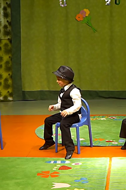 Children in a school setting, performing with formal attire on a stage decorated for spring or summer.
