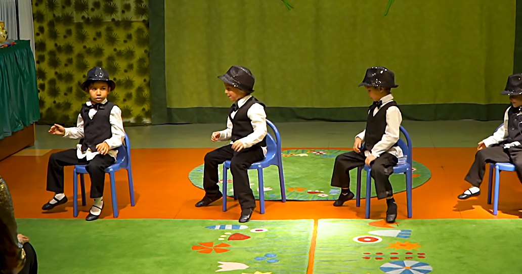 Children in a school setting, performing with formal attire on a stage decorated for spring or summer.