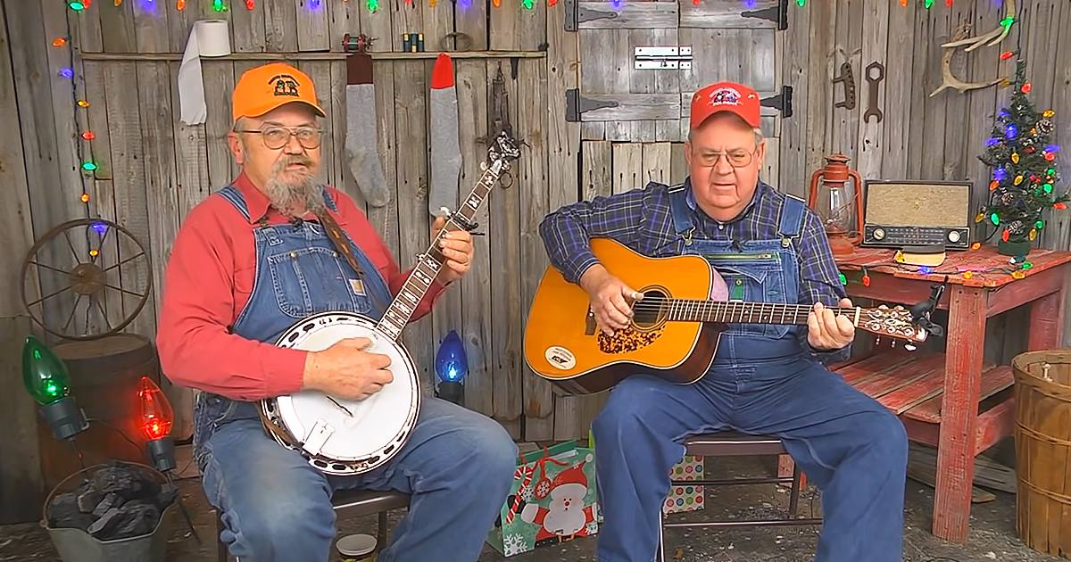 Two adult men playing musical instruments in a rustic cabin with Christmas lights, suggesting a holiday setting.