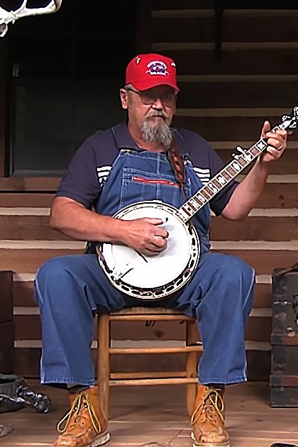 Two middle-aged to older adult males playing musical instruments in a rustic cabin porch setting, surrounded by wooden furniture and antler decorations.