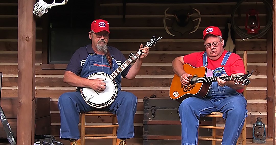 Two middle-aged to older adult males playing musical instruments in a rustic cabin porch setting, surrounded by wooden furniture and antler decorations.