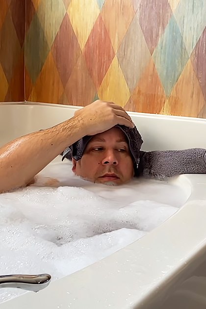 An adult male and his English Bulldog in a bathroom with colorful wallpaper, daytime lighting, and a white bathtub filled with soapy water.
