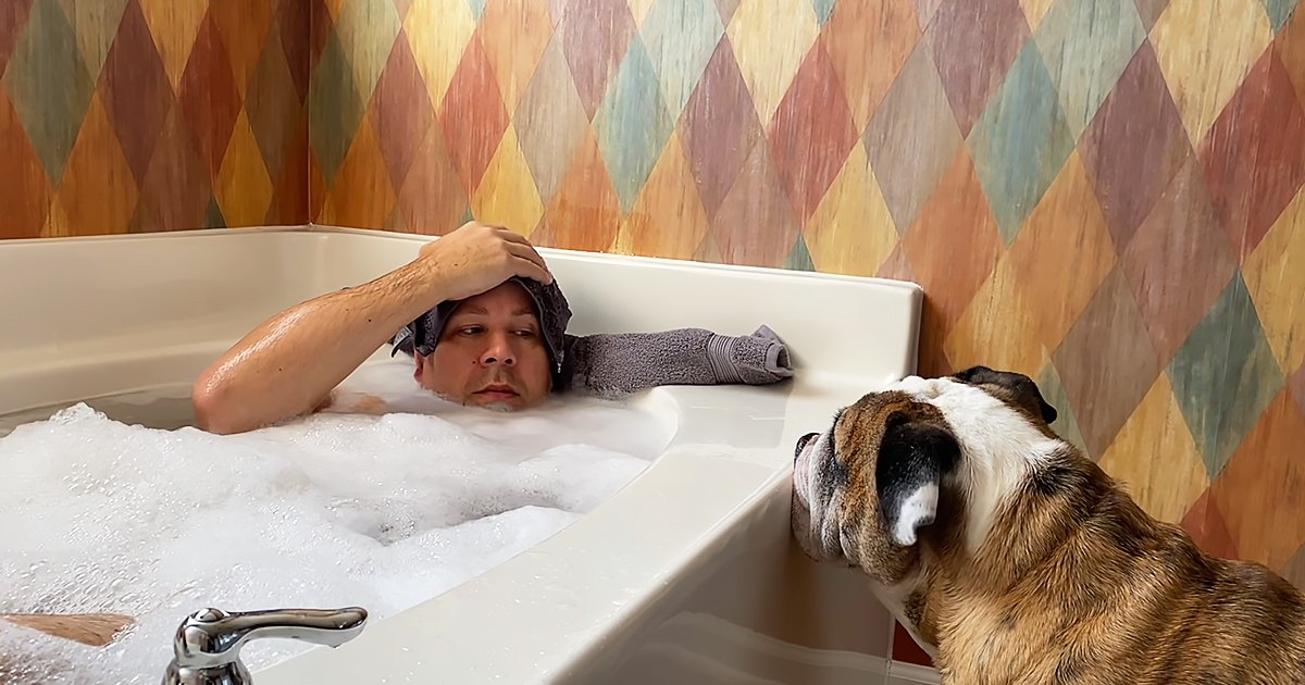 An adult male and his English Bulldog in a bathroom with colorful wallpaper, daytime lighting, and a white bathtub filled with soapy water.