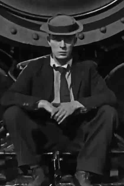 Adult male in dark suit, white shirt, dark tie and fedora hat seated on industrial machine in an old factory setting under natural daylight.