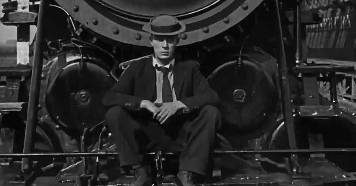 Adult male in dark suit, white shirt, dark tie and fedora hat seated on industrial machine in an old factory setting under natural daylight.
