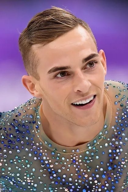 A male figure skater in a sparkling blue costume smiles while gliding on ice during an Olympic event.