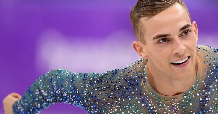 A male figure skater in a sparkling blue costume smiles while gliding on ice during an Olympic event.