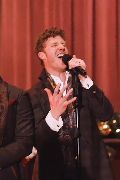 Group performance scene at an indoor event, possibly a concert or holiday celebration with formal attire and festive decor.