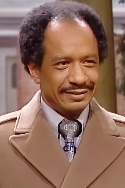 An adult male wearing a beige trench coat with a notched lapel, white collared shirt, and patterned tie stands indoors near an older adult male in a beige suit jacket. The scene includes traditional decor elements like a wooden door frame and suggests daytime lighting.