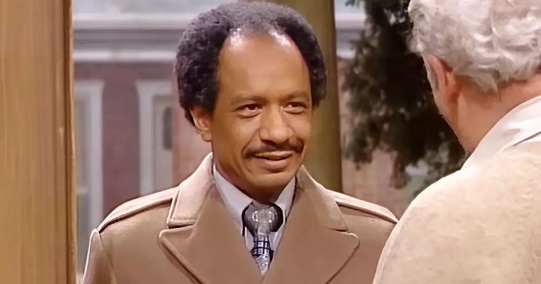 An adult male wearing a beige trench coat with a notched lapel, white collared shirt, and patterned tie stands indoors near an older adult male in a beige suit jacket. The scene includes traditional decor elements like a wooden door frame and suggests daytime lighting.