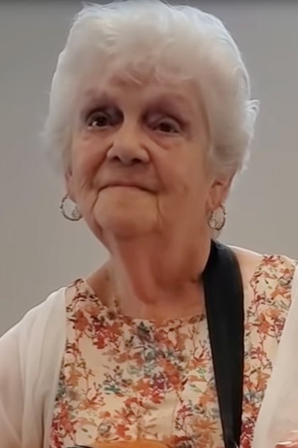 An older adult female wearing a floral-patterned top with a white cardigan or light jacket draped over her shoulders, holding a guitar strap around her neck in an indoor setting.