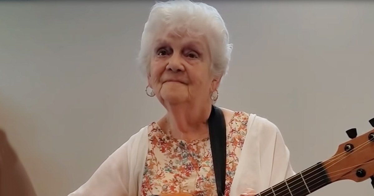 An older adult female wearing a floral-patterned top with a white cardigan or light jacket draped over her shoulders, holding a guitar strap around her neck in an indoor setting.