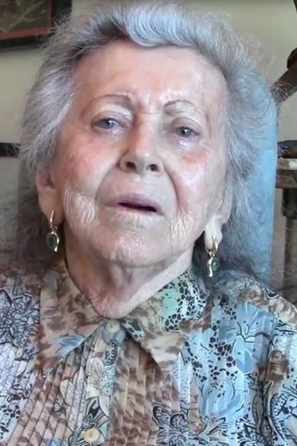 An older adult female in a home setting, wearing a patterned blouse with gold hoop earrings. A dark-colored chair and framed picture are visible in the background.