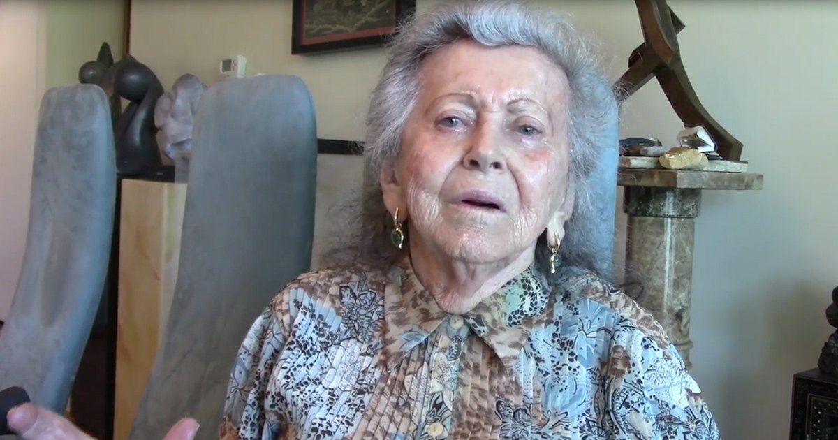 An older adult female in a home setting, wearing a patterned blouse with gold hoop earrings. A dark-colored chair and framed picture are visible in the background.