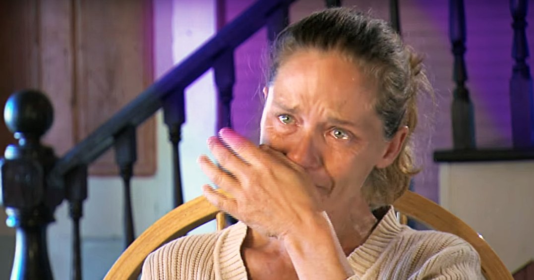 A middle-aged woman wearing a beige ribbed sweater in an indoor setting with a staircase and purple-painted walls, suggesting daytime.