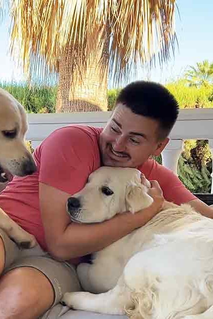 A man with two golden retriever dogs outdoors, wearing a red t-shirt and beige shorts. Palm fronds and greenery suggest spring or summer.