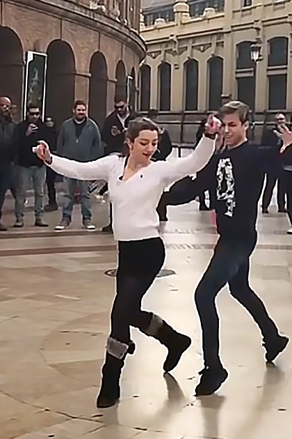 A couple dancing in a public square with historical architecture under soft evening light; people of various ages wearing winter clothing.