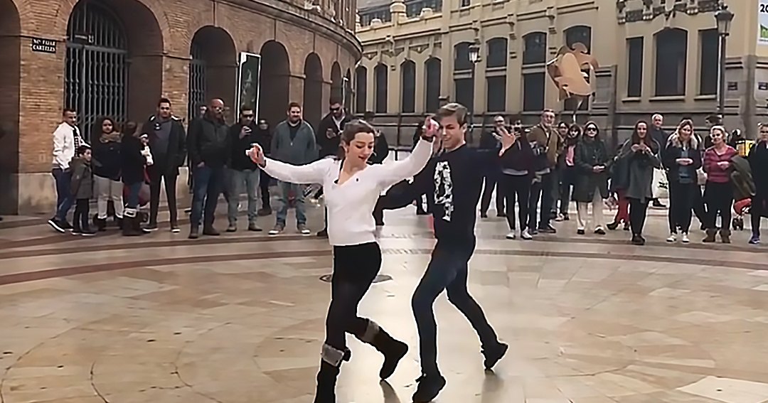 A couple dancing in a public square with historical architecture under soft evening light; people of various ages wearing winter clothing.
