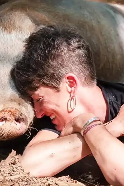 An adult female wearing a black tank top with colorful patterns, interacting with a potbellied pig in an outdoor farmyard setting during daylight.