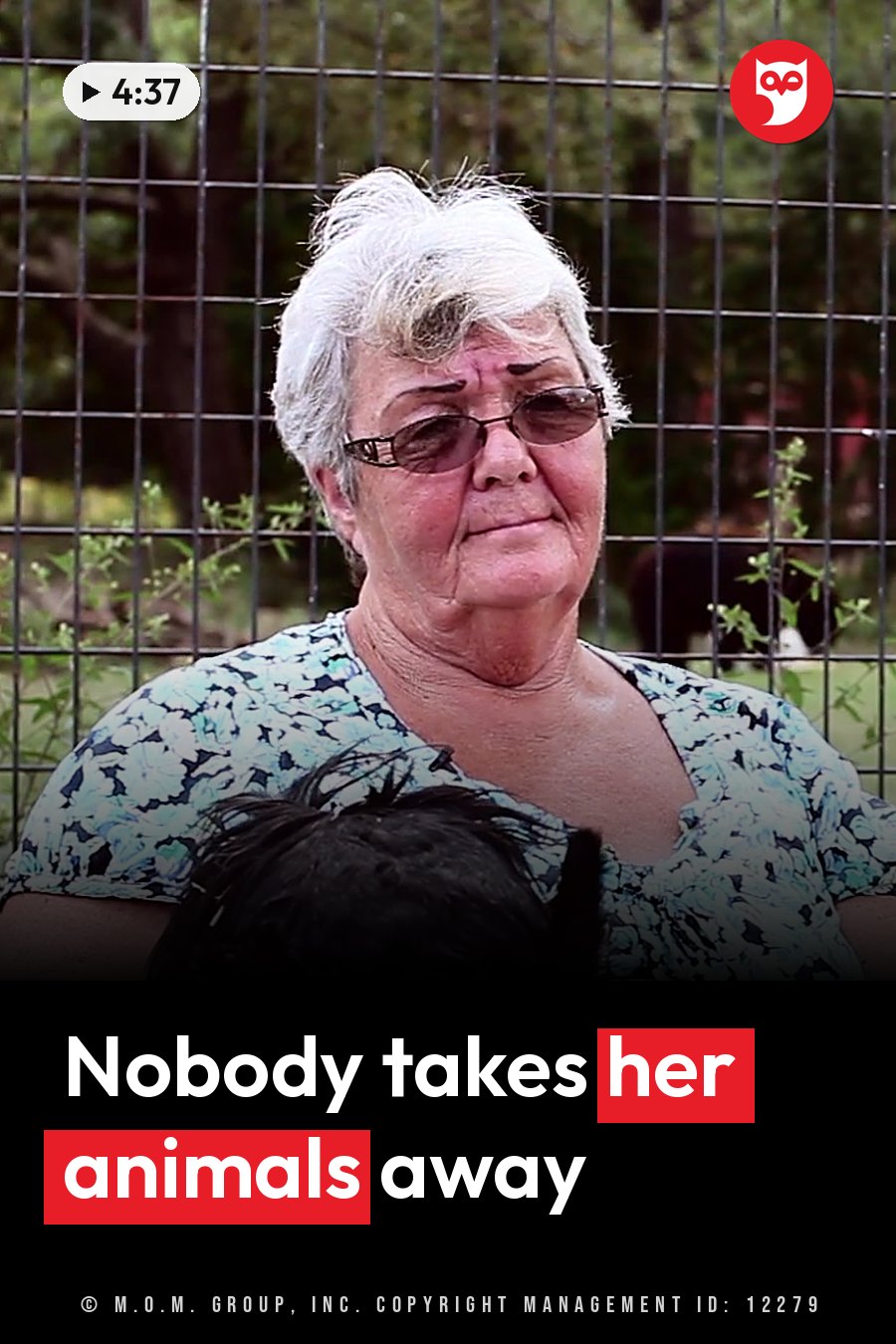 An older woman with short white hair and glasses holds a small black dog near a wire fence outdoors. A headline runs in white letters across the lower third.