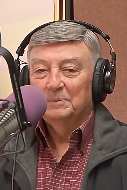 An older adult male wearing a dark gray jacket over a red and white checkered shirt, with large black over-ear headphones, sits in front of a microphone at '104.1 KRBE' radio station.