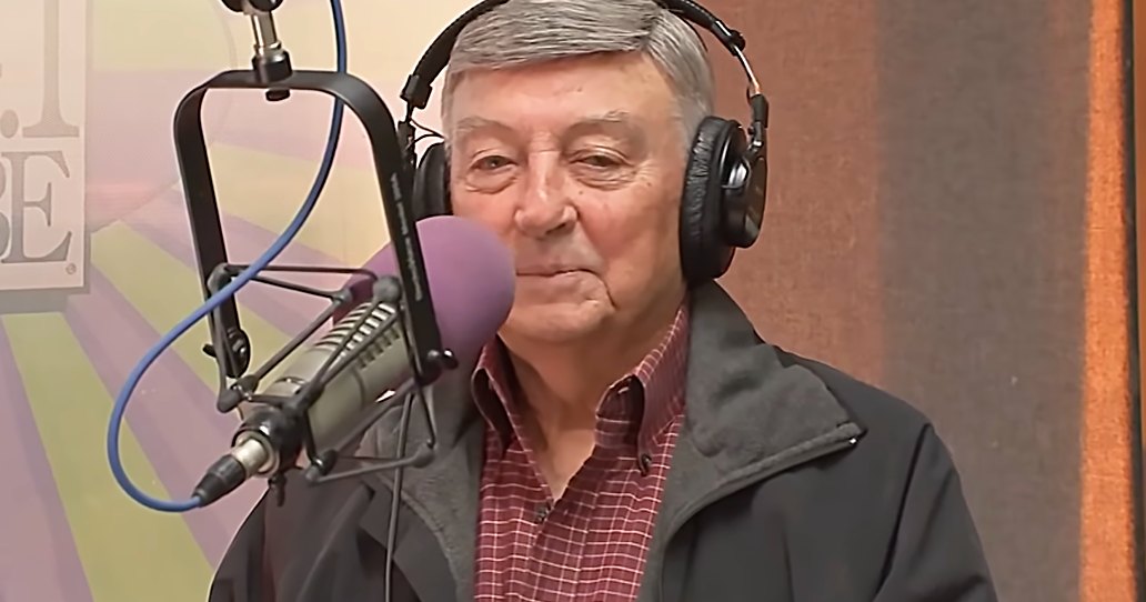 An older adult male wearing a dark gray jacket over a red and white checkered shirt, with large black over-ear headphones, sits in front of a microphone at '104.1 KRBE' radio station.