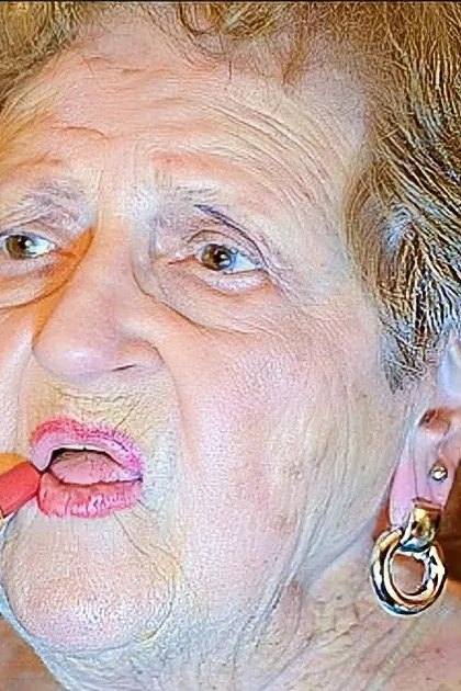 An older adult female in a home setting, wearing a dark green dress with intricate patterns and long sleeves. She is applying lipstick while holding the tube.