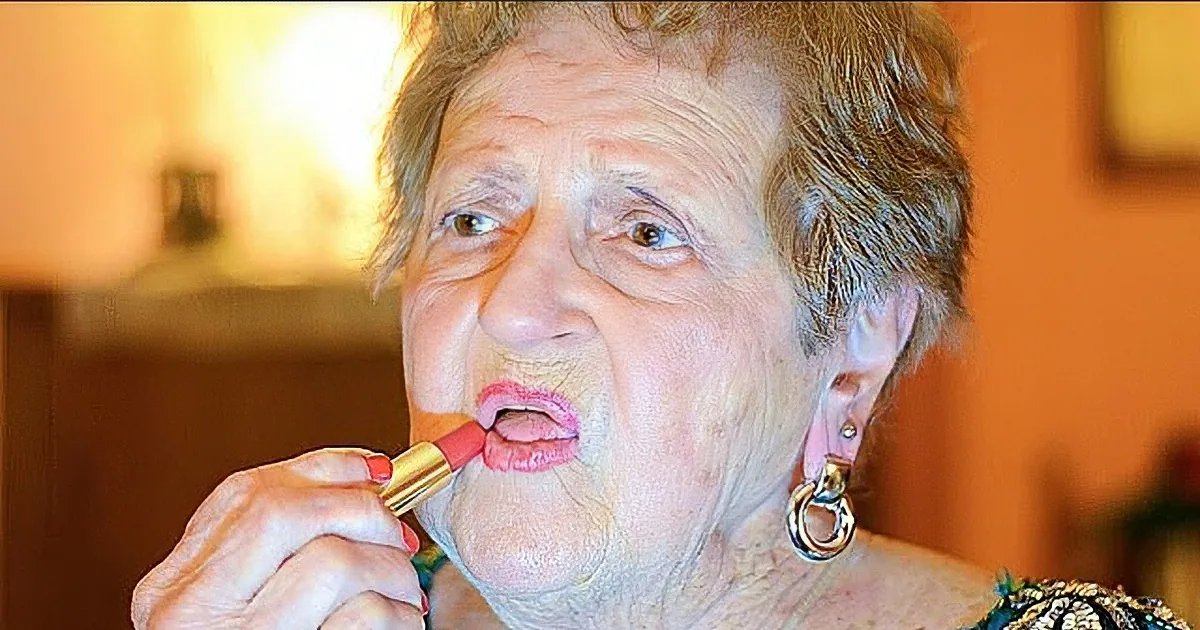An older adult female in a home setting, wearing a dark green dress with intricate patterns and long sleeves. She is applying lipstick while holding the tube.