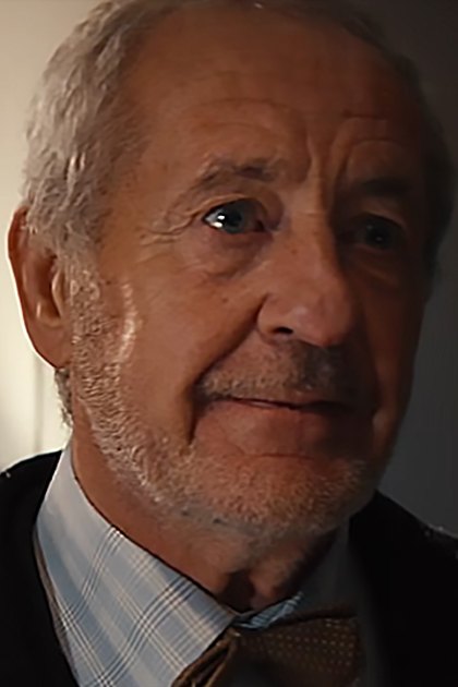 Older adult male wearing dark suit jacket with textured pattern over light blue and white checkered shirt, brown polka-dot tie; young adult male in dark suit jacket, white dress shirt, dark-colored tie or bowtie in a softly lit indoor setting with neutral walls.