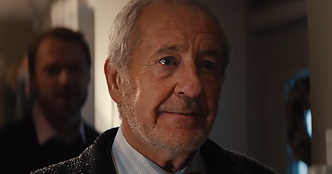Older adult male wearing dark suit jacket with textured pattern over light blue and white checkered shirt, brown polka-dot tie; young adult male in dark suit jacket, white dress shirt, dark-colored tie or bowtie in a softly lit indoor setting with neutral walls.