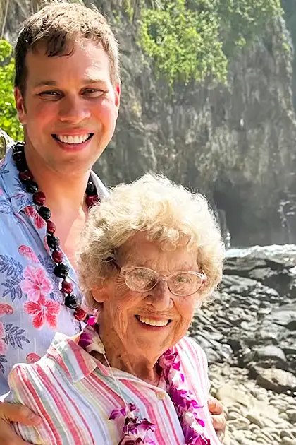 Young adult male in a light blue Hawaiian shirt with pink floral patterns, older adult female in striped shirt by rocky coastline at daytime tropical location.