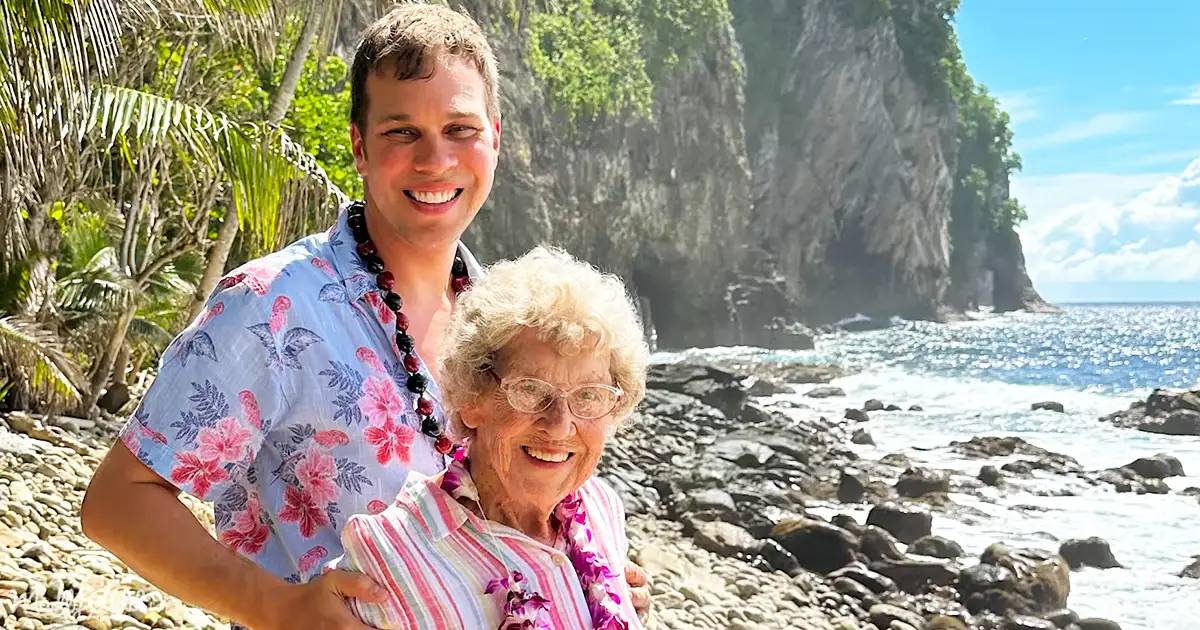 Young adult male in a light blue Hawaiian shirt with pink floral patterns, older adult female in striped shirt by rocky coastline at daytime tropical location.