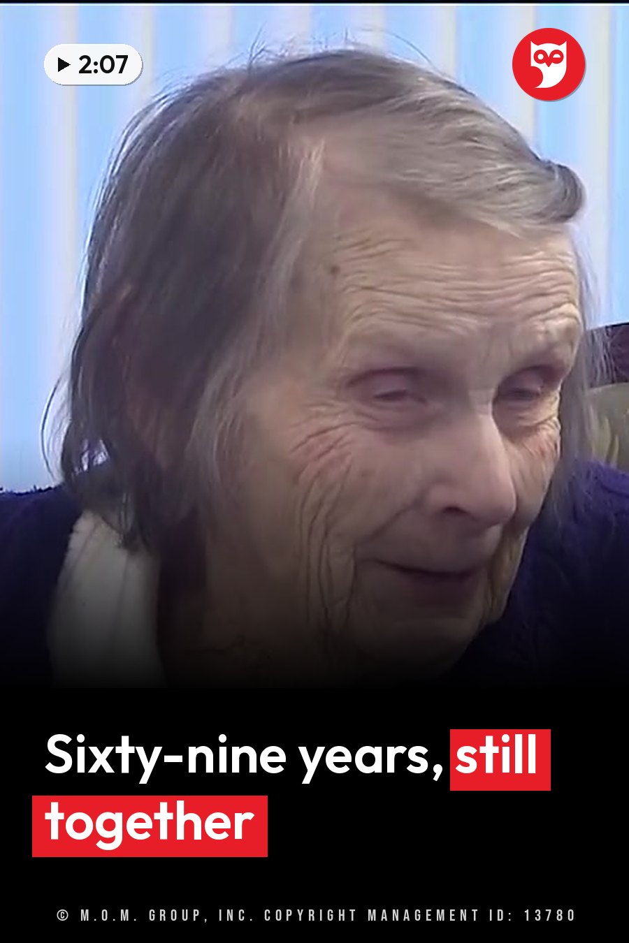 An elderly woman with gray hair and a purple top looks down while seated indoors near blue curtains. A headline runs in white letters across the lower third.