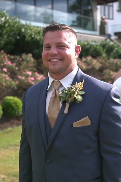 A wedding couple, a male in a dark blue formal suit with a light brown tie and pocket square, and a female in a strapless white wedding gown with a floral headpiece, standing outdoors at what appears to be a wedding venue.