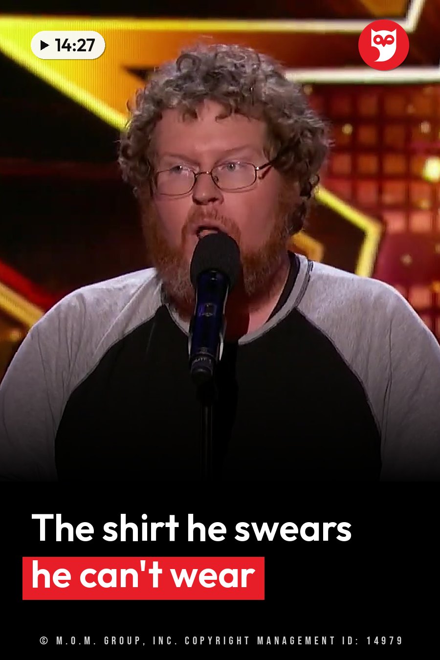 A man with curly hair, glasses and a beard holds a microphone to his mouth on a brightly lit stage. A headline runs in white letters across the lower third.