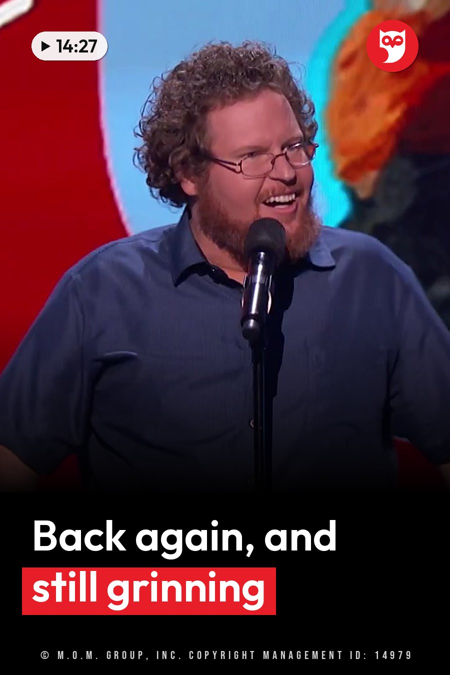 A man with curly hair and glasses smiles as he speaks into a microphone on a stage. A headline runs in white letters across the lower third.