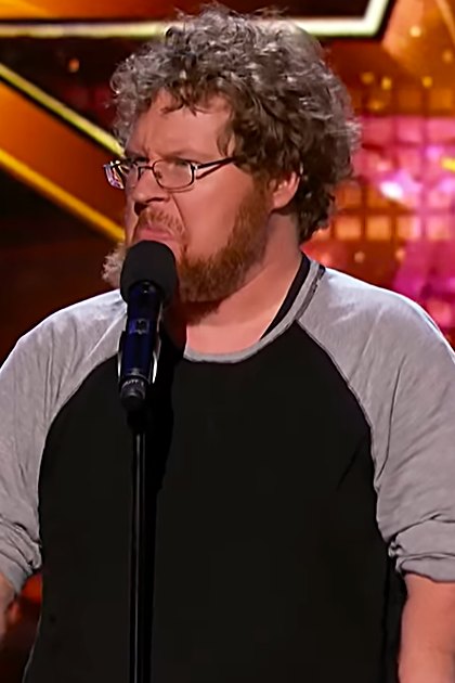 A middle-aged adult male wearing a long-sleeved raglan shirt with a relaxed fit and crew neck, performing on stage at an entertainment competition show.