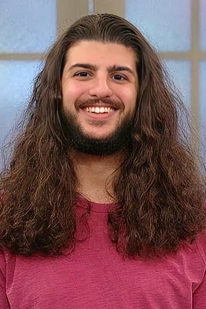 A man with long hair and a beard smiles while wearing a pink shirt indoors near a window.