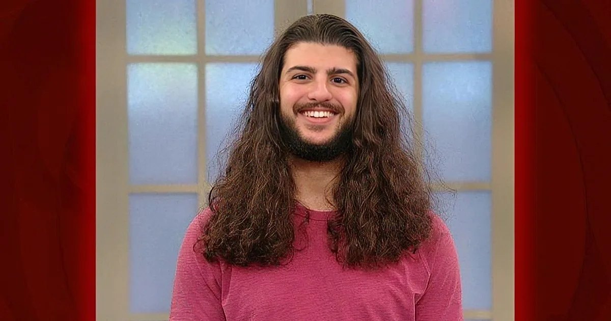 A man with long hair and a beard smiles while wearing a pink shirt indoors near a window.