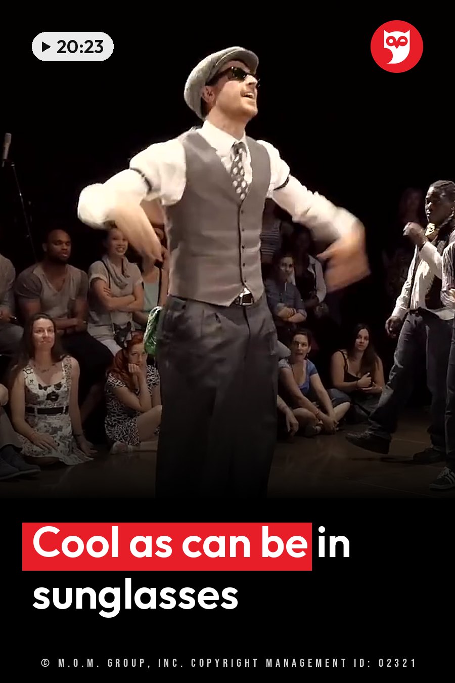 A man in a vest, hat and sunglasses dances on a stage in front of seated audience members. A headline runs in white letters across the lower third.