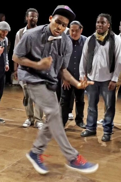 A young adult male dancer in an indoor dance studio or performance space, surrounded by various individuals engaged in different activities.