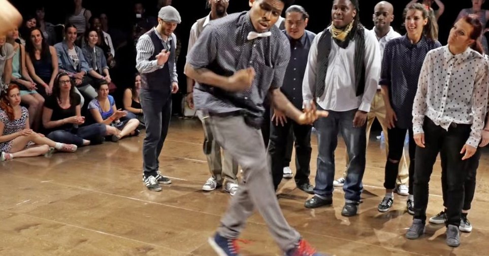 A young adult male dancer in an indoor dance studio or performance space, surrounded by various individuals engaged in different activities.