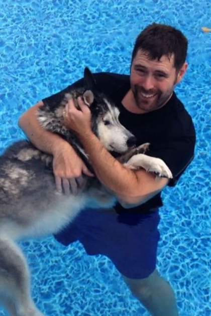 An adult male holding a gray, white, and black husky in a swimming pool setting