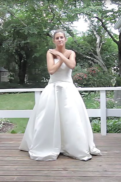 A young adult female in a modern white wedding dress stands on a wooden deck outside her house during spring or summer with soft evening light.