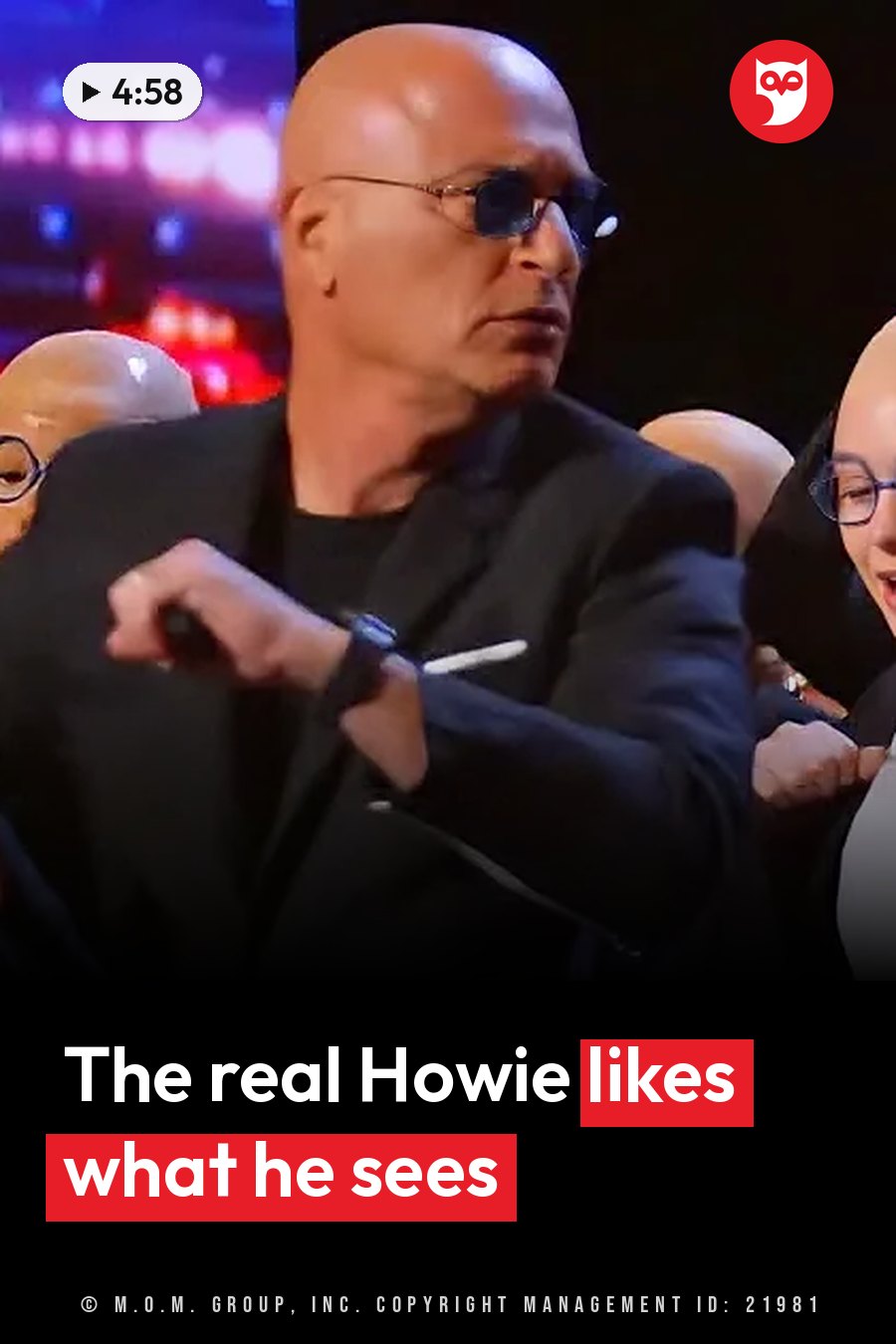 A bald man in sunglasses and a black suit sits and points forward with his right hand. A headline runs in white letters across the lower third.