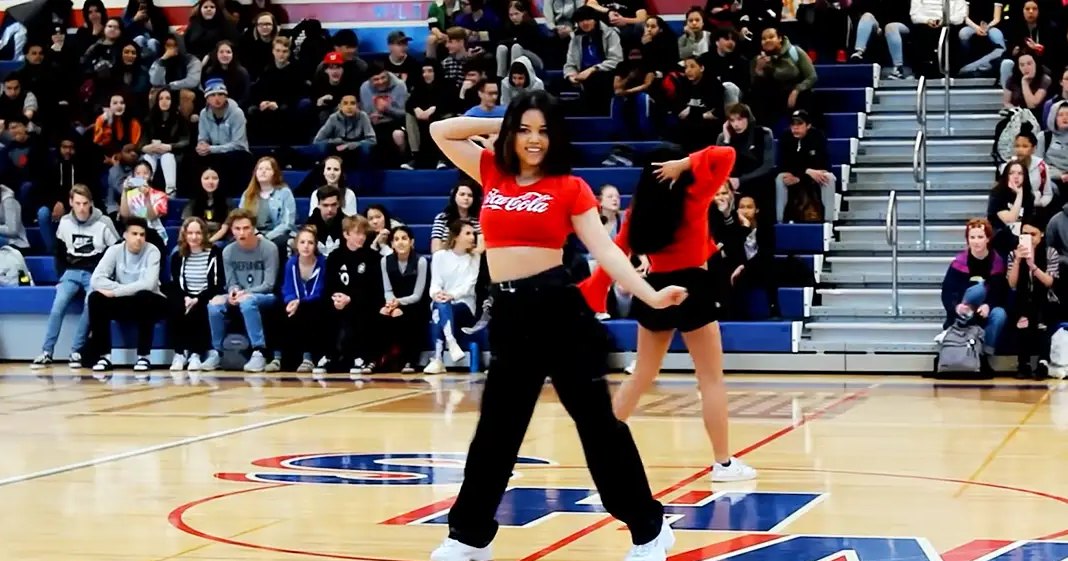 Young adult female dancers practicing in a gymnasium with an engaged audience wearing casual attire.