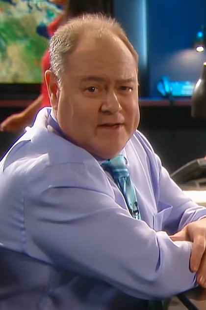 A middle-aged man wearing a light purple patterned dress shirt, a wristwatch, and standing in an office setting with another partially visible person behind him.