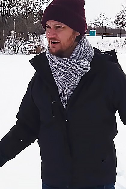 A young adult male wearing a dark jacket and scarf in a snowy outdoor setting during winter.