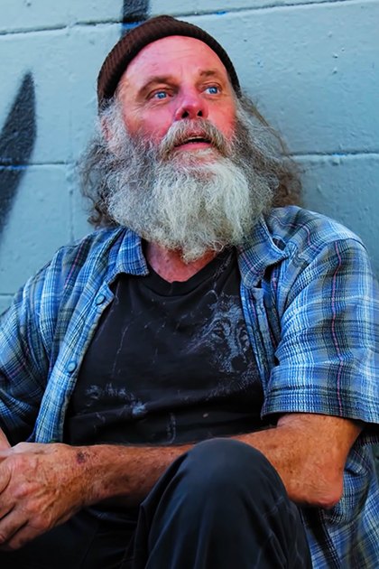 Older adult male wearing plaid short-sleeved shirt, dark t-shirt, black pants, and beanie hat, holding disposable coffee cup in an urban setting with graffiti on a blue brick wall during daytime.